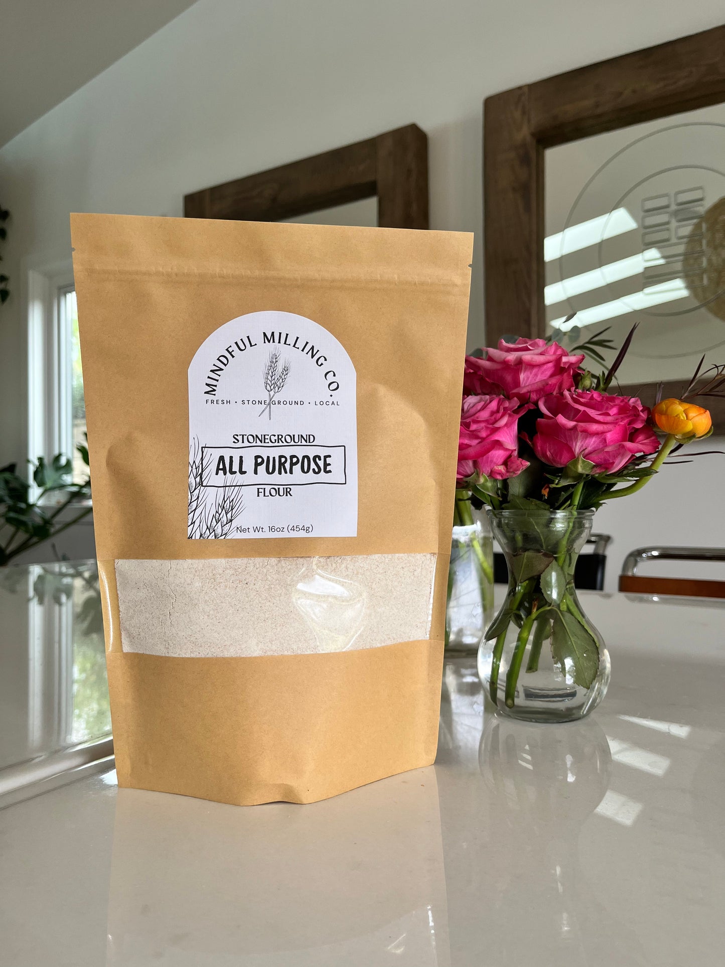 A bag of fresh milled flour on a white table with roses and a mirror in the background; brown kraft paper zip top pouch with a narrow rectangular window that spans with width of the bag, about a third of the way from the bottom. A textured white arched label with the Mindful Milling Co logo and the words “stoneground all purpose flour” with a line art wheat stalk, the weight measurement of the bag (1lb/454g).