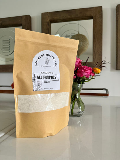 A bag of fresh milled flour on a white table with roses and a mirror in the background; brown kraft paper zip top pouch with a narrow rectangular window that spans with width of the bag, about a third of the way from the bottom. A textured white arched label with the Mindful Milling Co logo and the words “stoneground all purpose flour” with a line art wheat stalk, the weight measurement of the bag (1lb/454g).