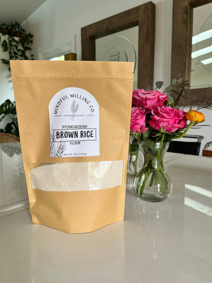 A bag of fresh milled flour on a white table with roses and a mirror in the background; brown kraft paper zip top pouch with a narrow rectangular window that spans with width of the bag, about a third of the way from the bottom. A textured white arched label with the Mindful Milling Co logo and the words “stoneground brown rice flour” with a line art wheat stalk, the weight measurement of the bag (1lb/454g). 